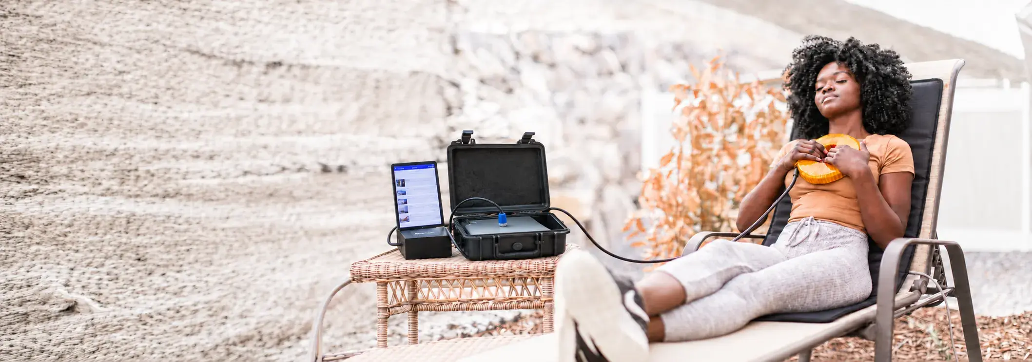 A woman relaxing outdoors in a lounge chair holding the AmpCoil coil, with the AmpCoil system and tablet on the table beside her